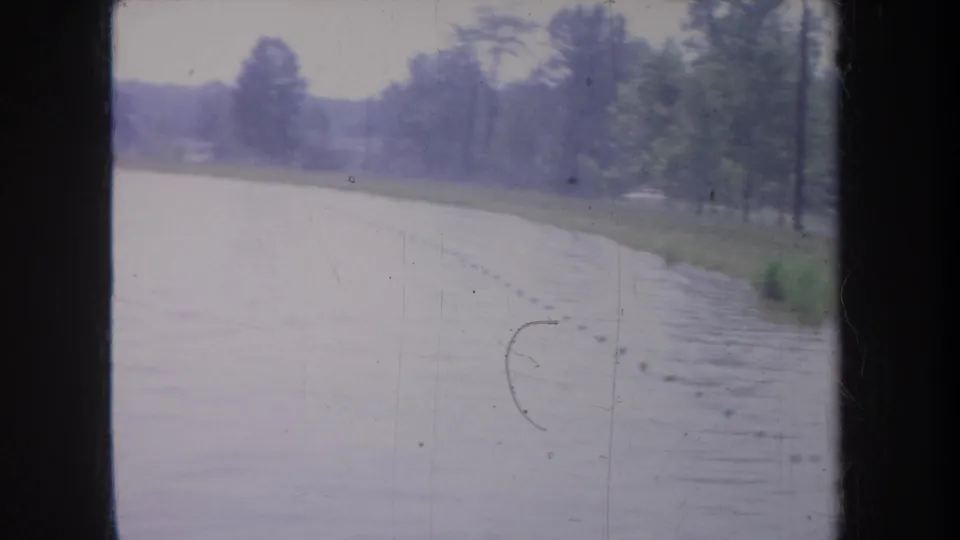 1968 Michigan home-movie view of a flooded lakeside or roadside area, with trees, grass, and a dark vertical window frame crossing the scene.