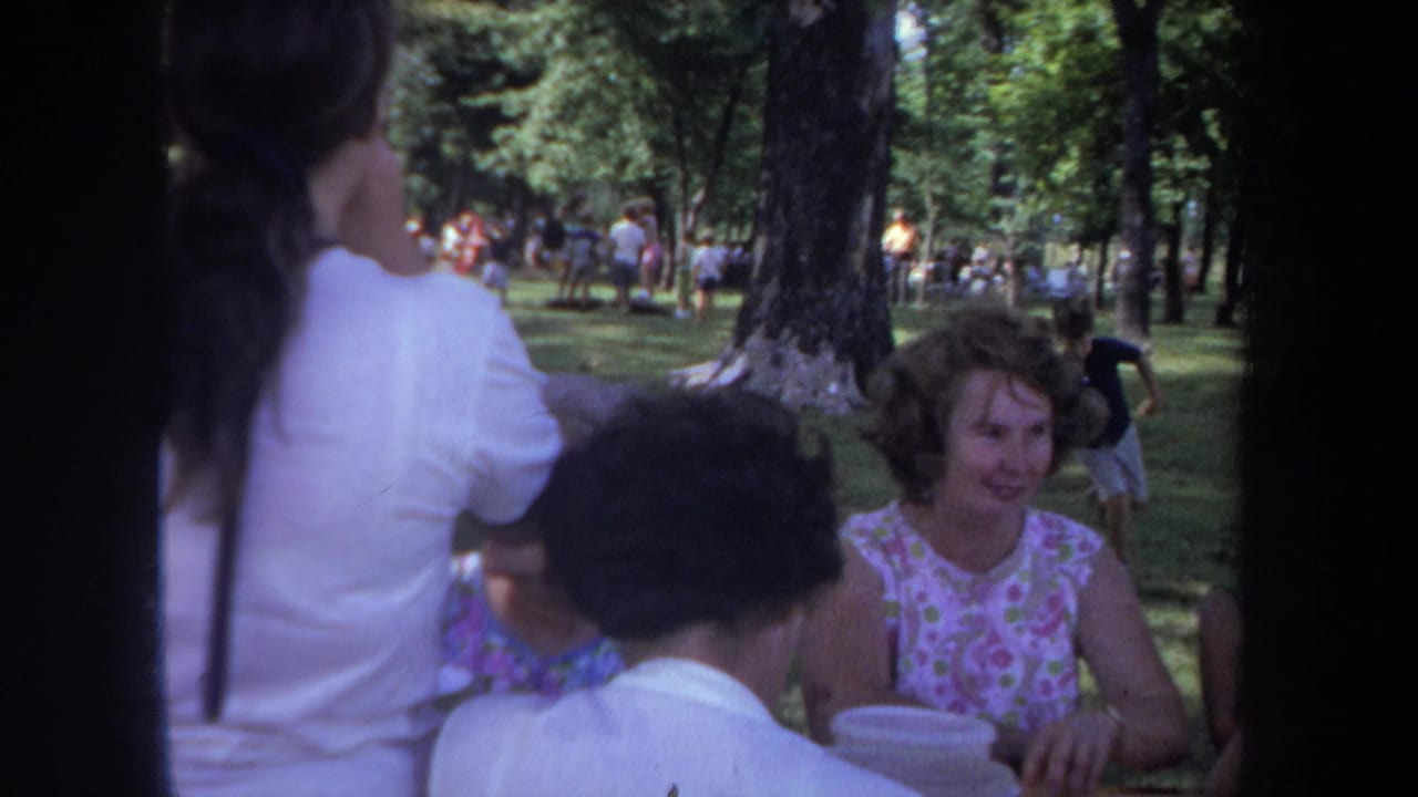 1968 Michigan 8mm frame of adult women at a park picnic table, partly obscured by the camera edge and nearby figures.
