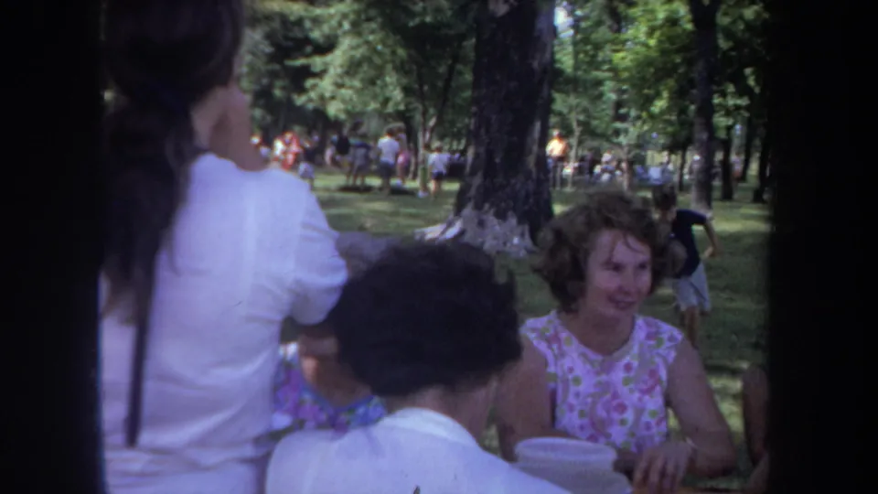 1968 Michigan 8mm frame of adult women at a park picnic table, partly obscured by the camera edge and nearby figures.