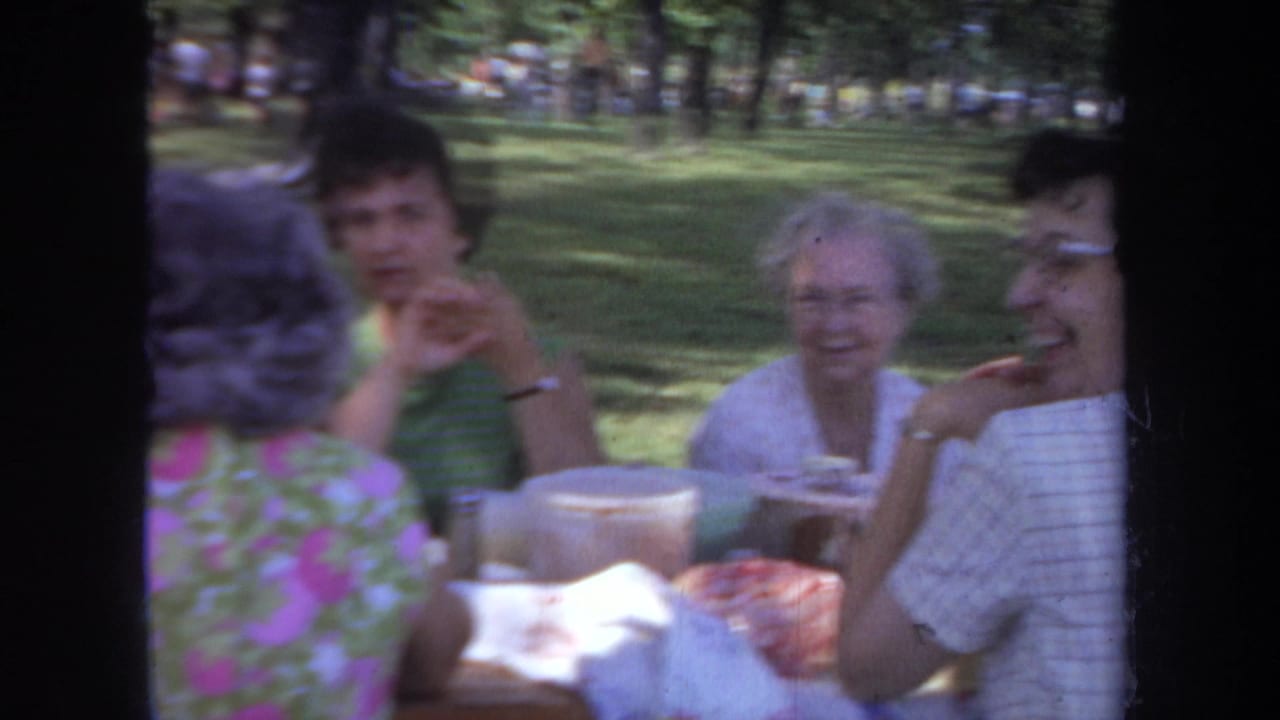 1968 Michigan home-movie frame of adult women in floral and sleeveless clothing seated at a park picnic table.