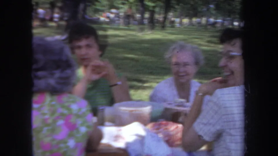 1968 Michigan home-movie frame of adult women in floral and sleeveless clothing seated at a park picnic table.