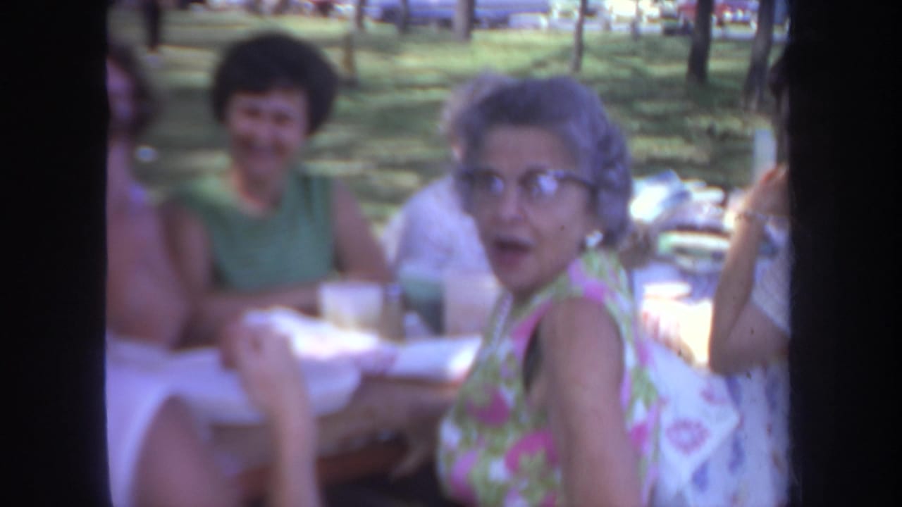 1968 Michigan home-movie frame with an older woman in glasses looking toward the camera during a park picnic.