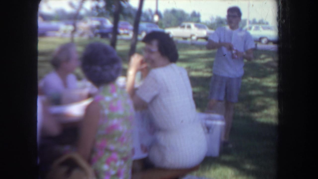 Several adults gather at a picnic table as a man in a white shirt and shorts stands nearby among trees and parked cars.