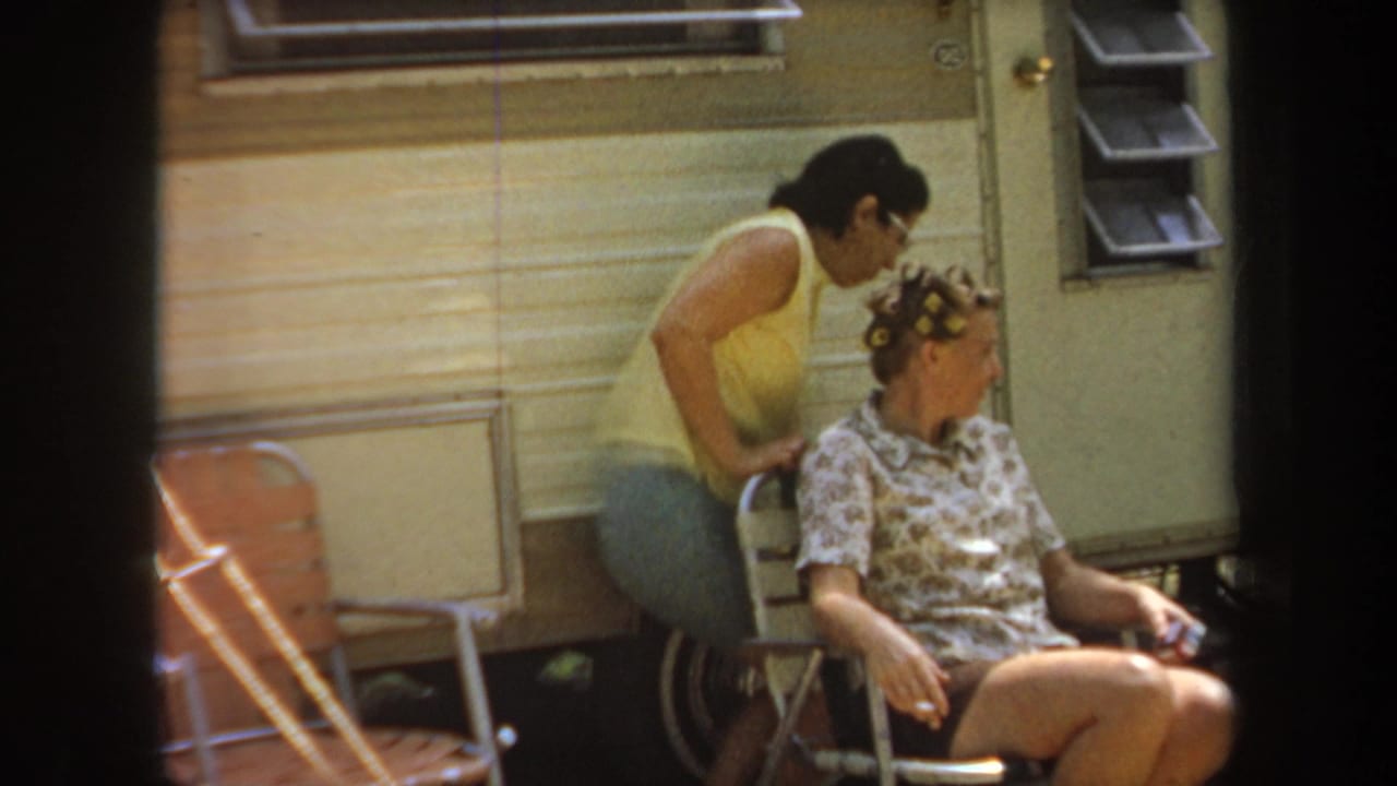 1968 Michigan home-movie frame showing adults beside a cream trailer, with a woman in curlers seated in a folding chair.