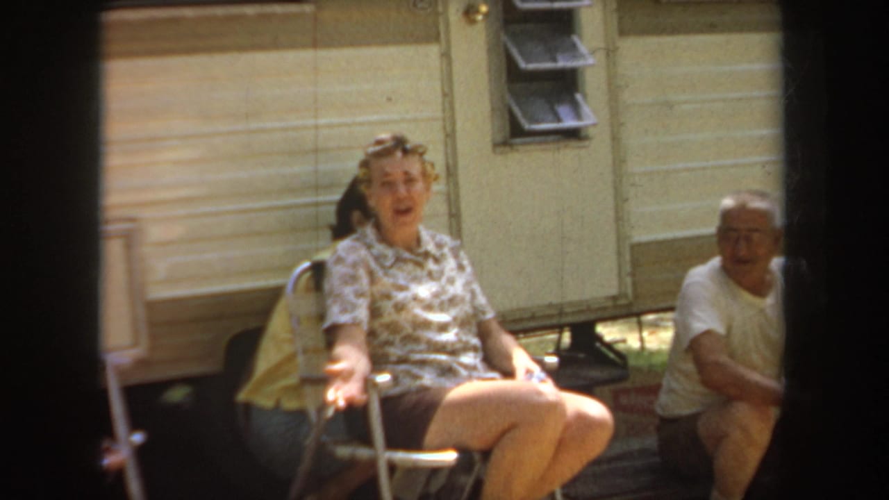 Woman in curlers gestures from a folding chair as a man sits beside her outside the trailer.