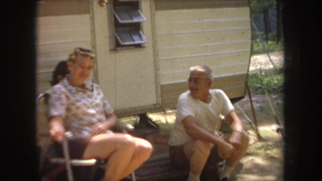 1968 Michigan home-movie frame showing two adults relaxing beside a travel trailer with trees in the background.