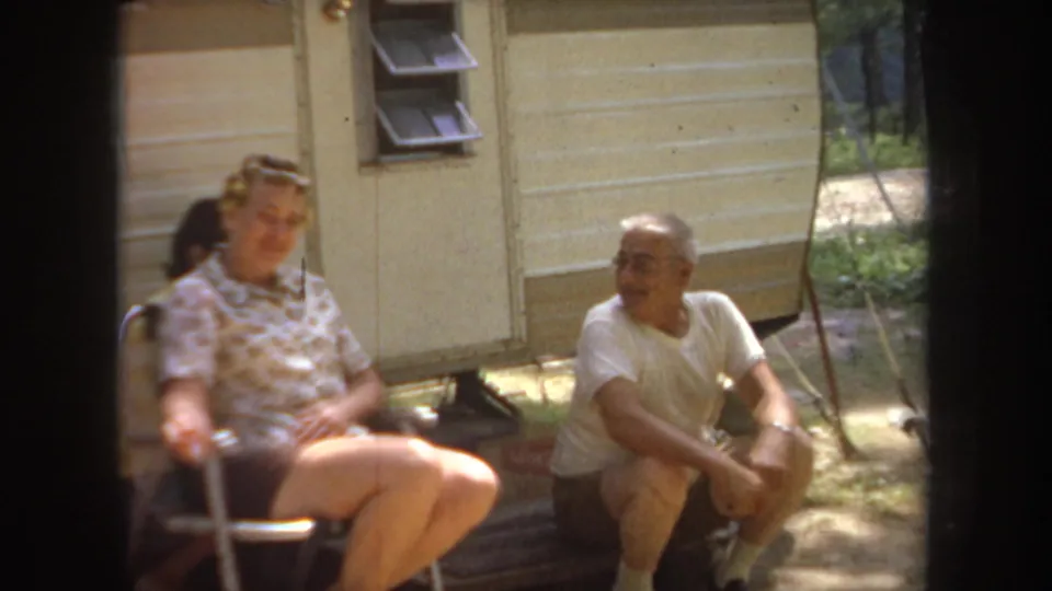1968 Michigan home-movie frame showing two adults relaxing beside a travel trailer with trees in the background.