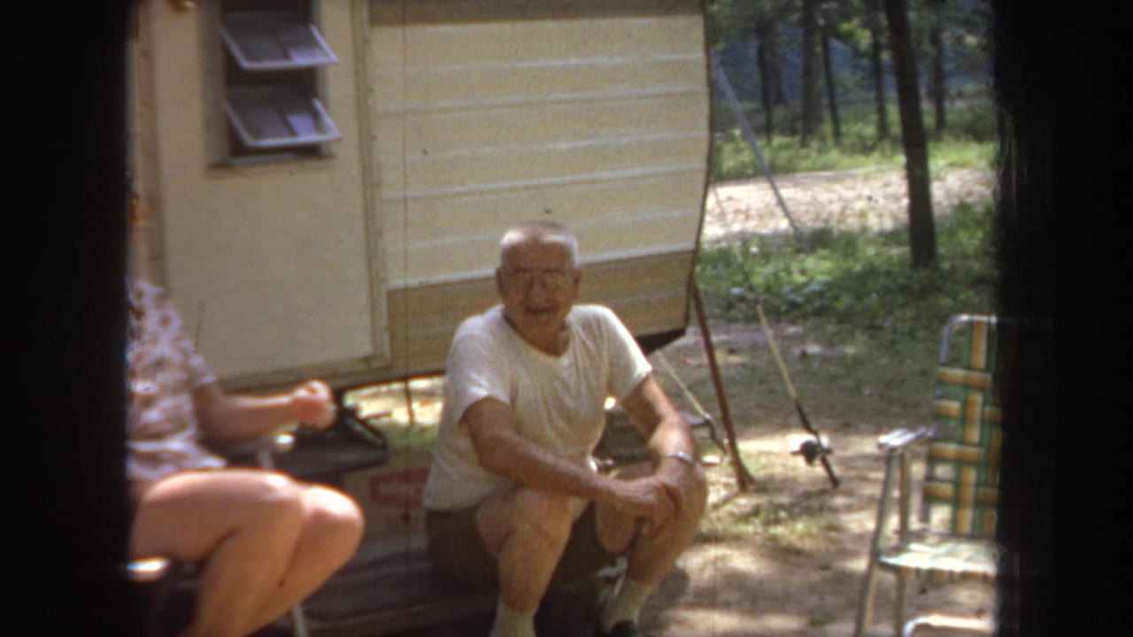 1968 Michigan frame of a seated man beside a cream travel trailer, with folding chairs and wooded ground visible.