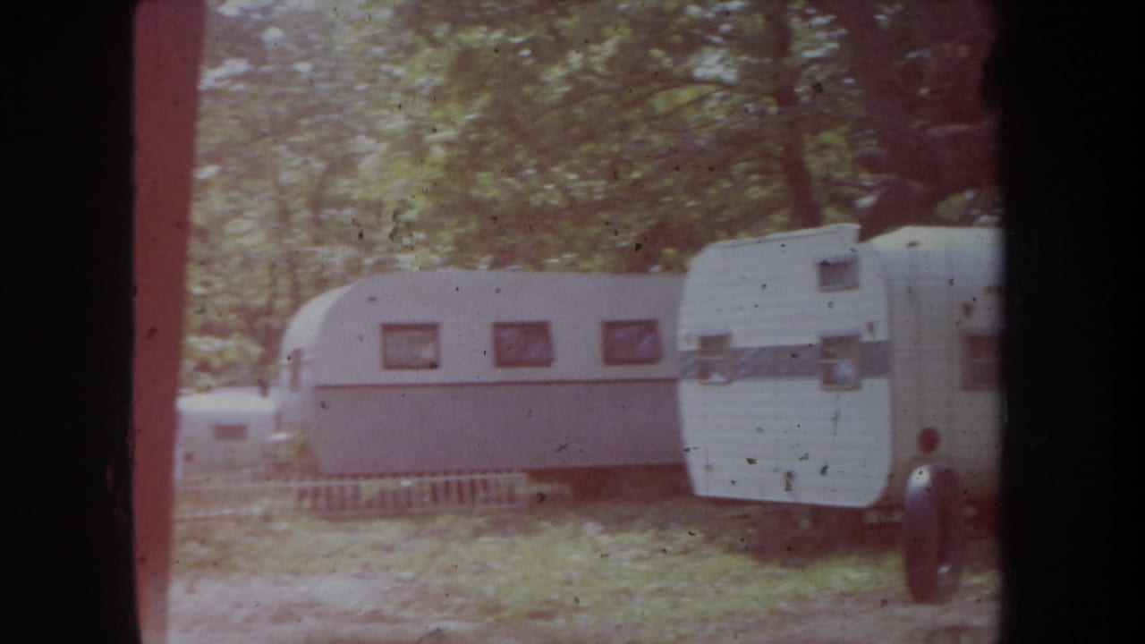 1968 Michigan home-movie frame showing white travel trailers lined up in a shaded forest campground.