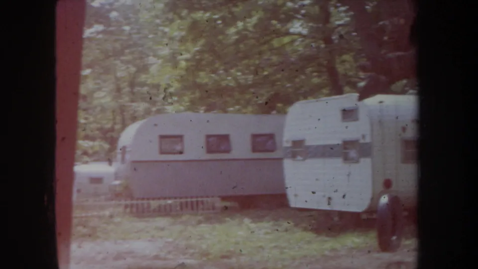 1968 Michigan home-movie frame showing white travel trailers lined up in a shaded forest campground.