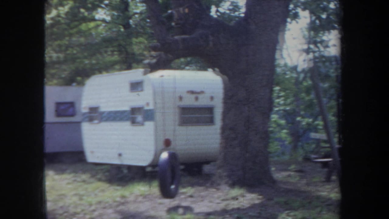 1968 Michigan campground view with white travel trailers, a large tree trunk, and wooded shade.