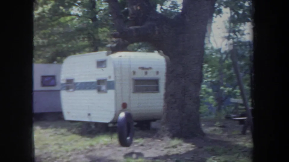 1968 Michigan campground view with white travel trailers, a large tree trunk, and wooded shade.