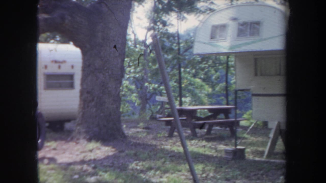 1968 Michigan home-movie frame showing a raised white trailer with a green stripe beside a picnic table.