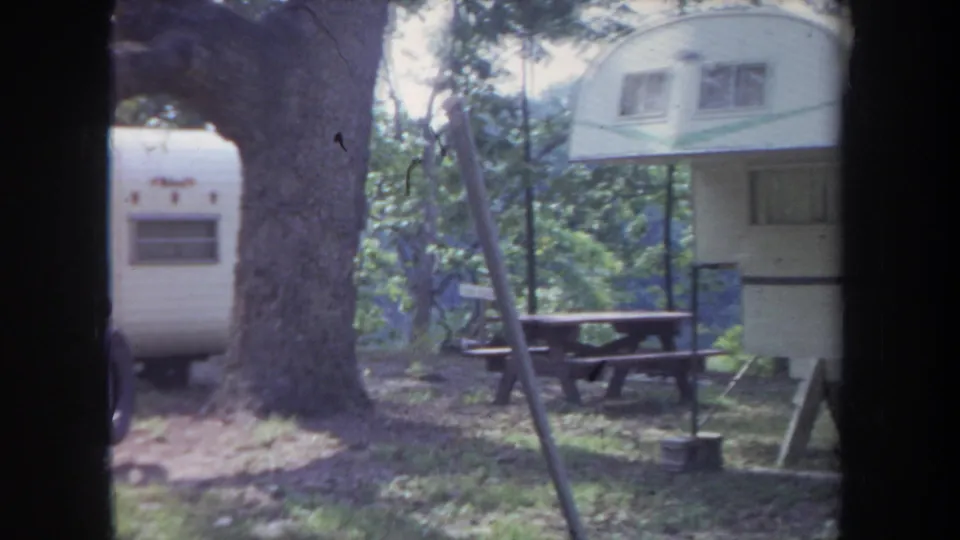 1968 Michigan home-movie frame showing a raised white trailer with a green stripe beside a picnic table.