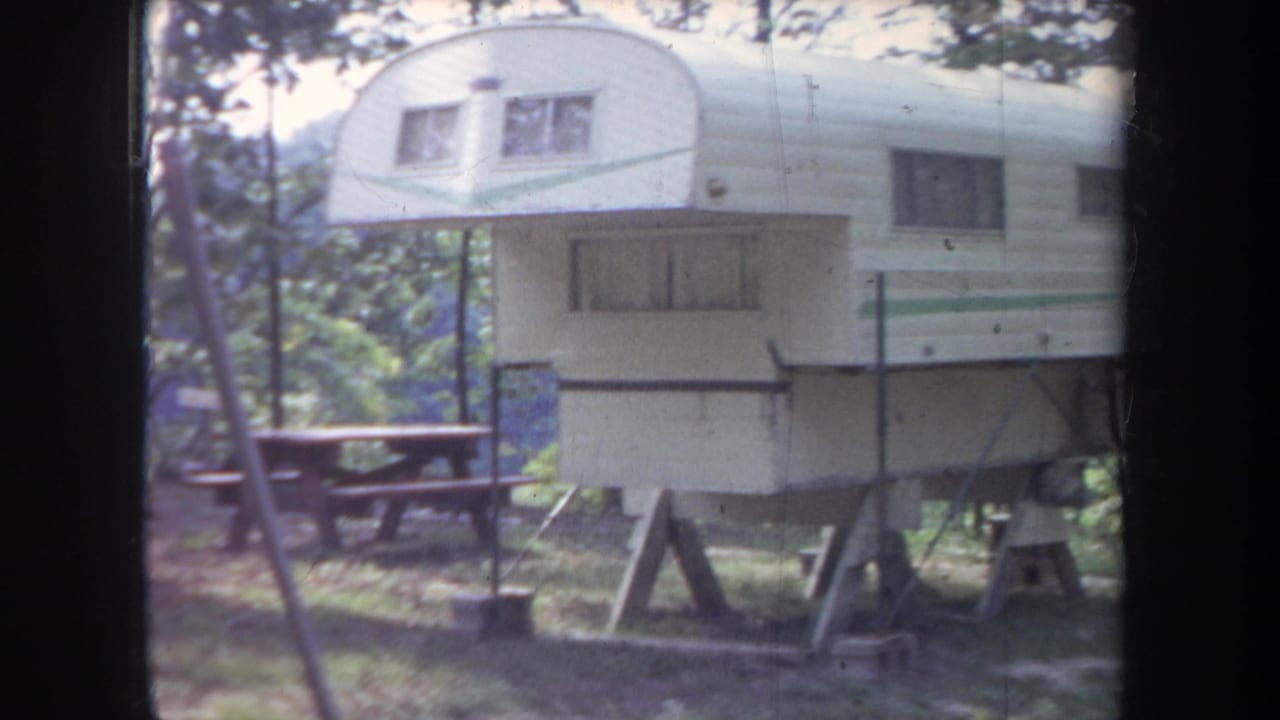 1968 Michigan frame showing a white camper trailer with green stripe, raised supports, and a picnic table in the woods.