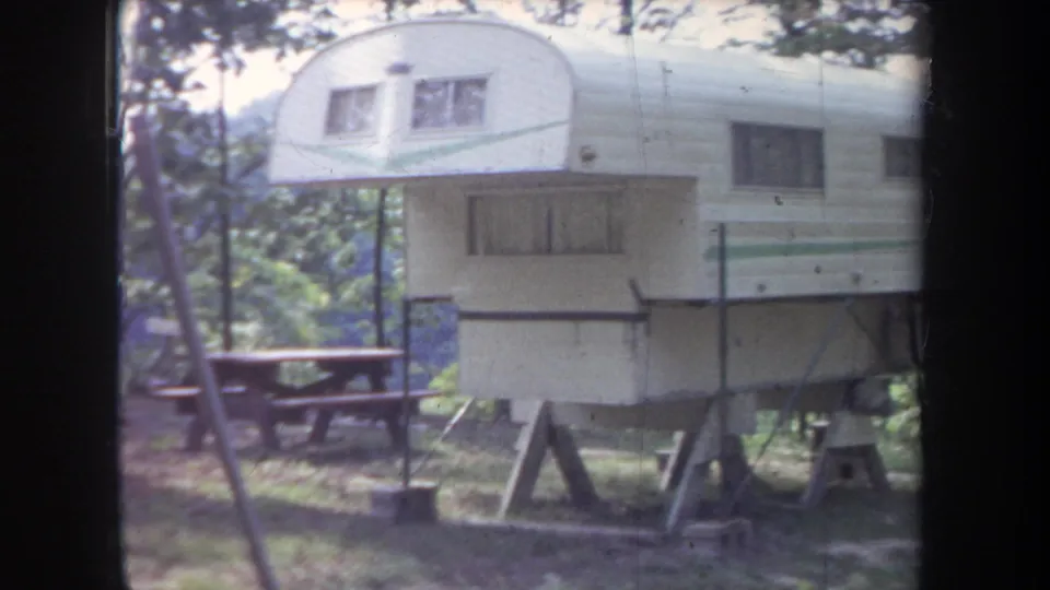 1968 Michigan frame showing a white camper trailer with green stripe, raised supports, and a picnic table in the woods.