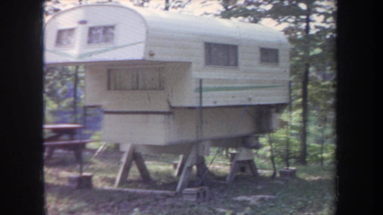 1968 Michigan 8mm frame showing a white-and-green camper trailer on wooden supports among trees at a campsite.