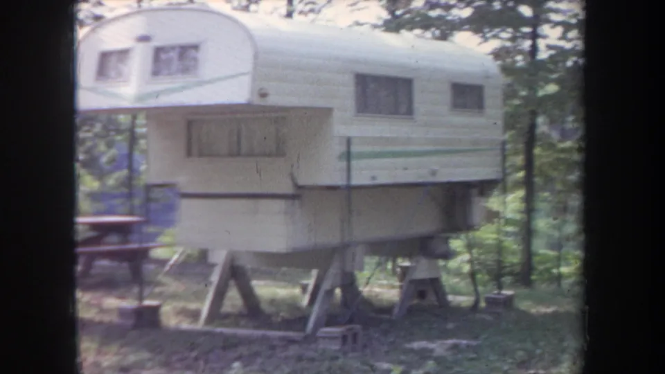 1968 Michigan 8mm frame showing a white-and-green camper trailer on wooden supports among trees at a campsite.