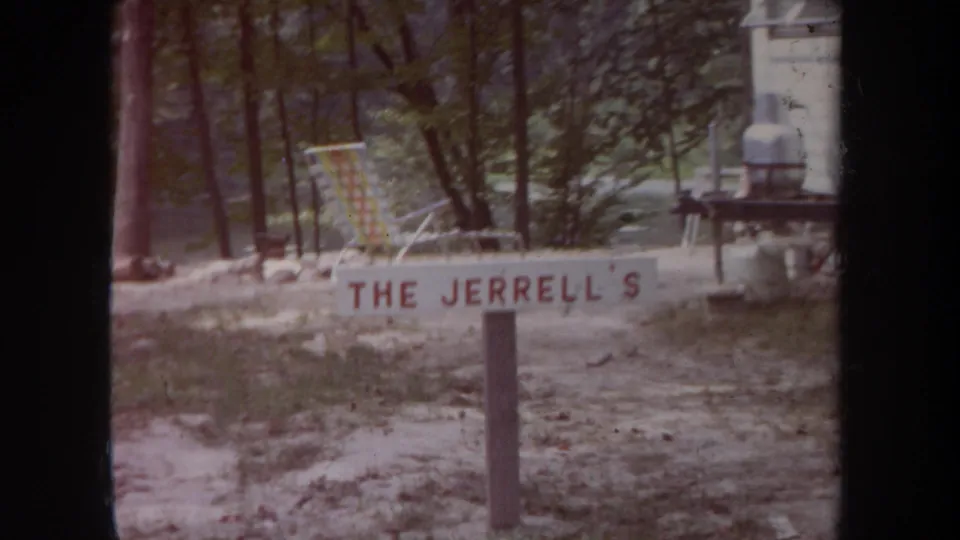 1968 Michigan campsite frame with a painted THE JERRELL'S sign, folding chair, small table, and camper edge in view.