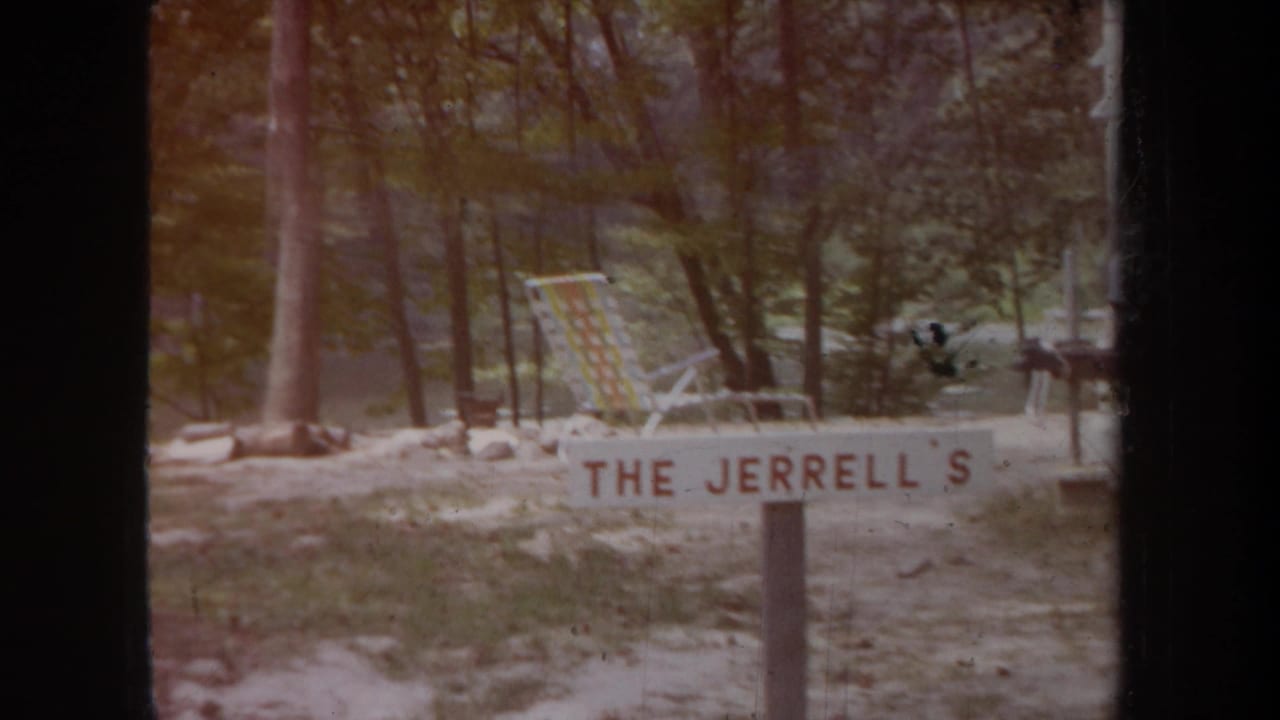 1968 Michigan home-movie frame showing a family-name campsite sign, folding lounge chair, trees, and sandy ground.