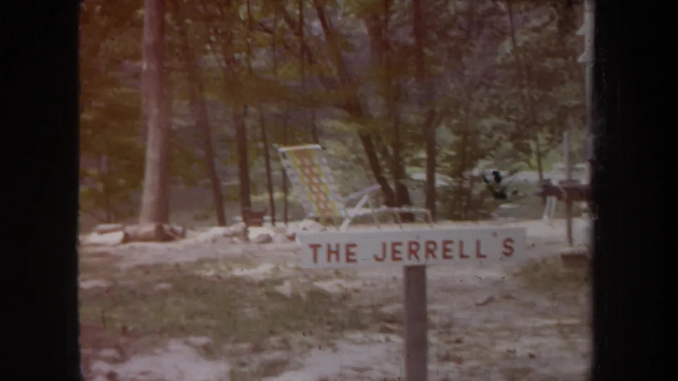 1968 Michigan home-movie frame showing a family-name campsite sign, folding lounge chair, trees, and sandy ground.