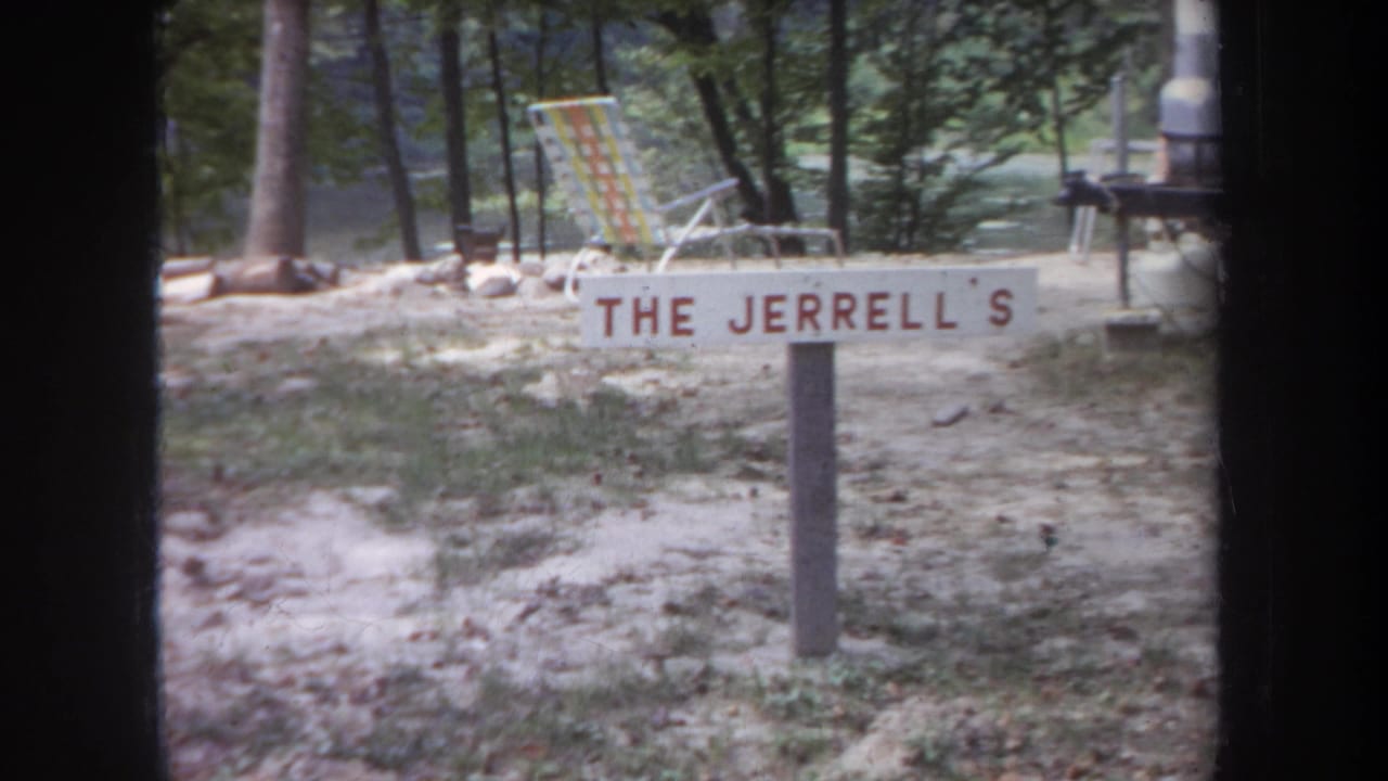 The Jerrell family sign stands in a wooded Michigan campsite with a striped chair and lake-area shade behind it.
