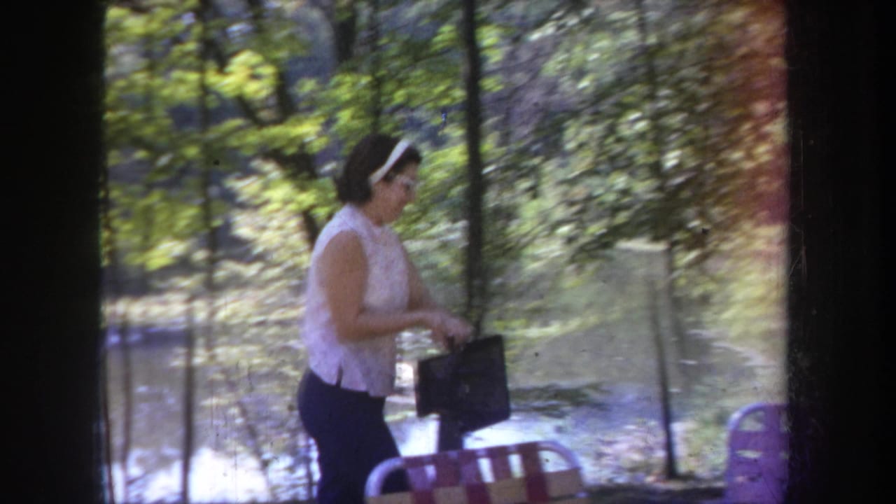 In 1968 Michigan home-movie footage, a woman stands near a lake with trees, a folding chair, and a dark bag.