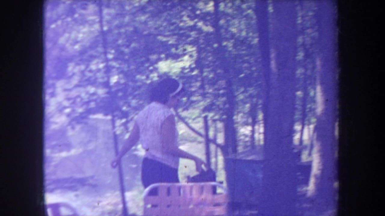 Purple-tinted frame of the woman walking near trees and campsite furniture.