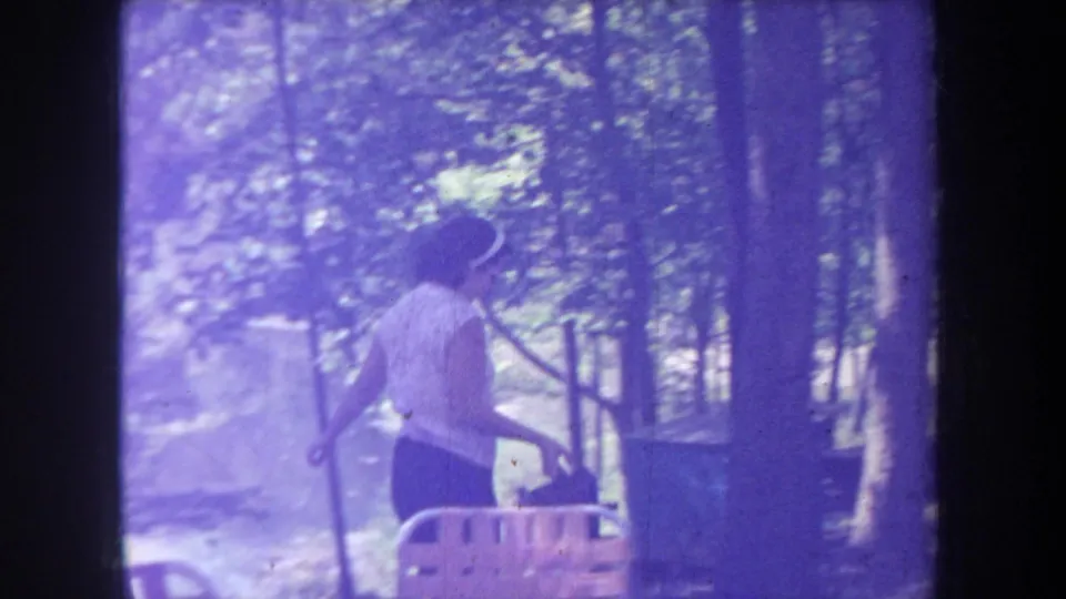Purple-tinted 1968 Michigan frame shows the woman moving through a wooded lakeside campsite with chairs nearby.