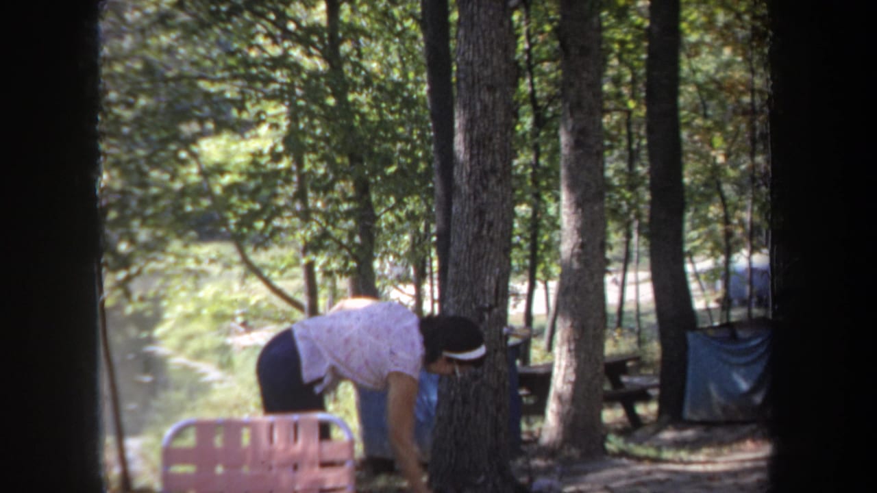 The woman bends toward the ground near a tree at the Michigan lakeside campsite, with water and picnic furniture visible.