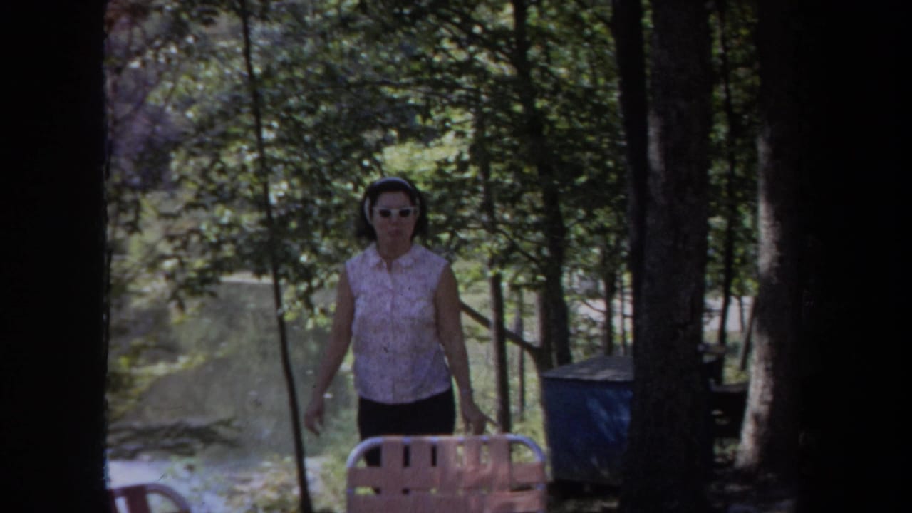 In 1968 Michigan home-movie footage, a woman in white sunglasses walks toward the camera among trees near outdoor seating.