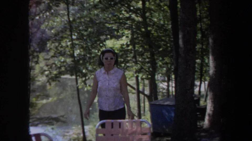 In 1968 Michigan home-movie footage, a woman in white sunglasses walks toward the camera among trees near outdoor seating.