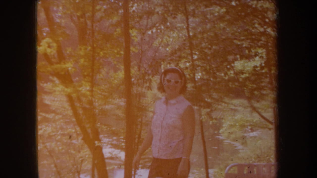 A warm color-shifted 8mm frame shows the woman in sunglasses paused among trees in the Michigan outdoor setting.