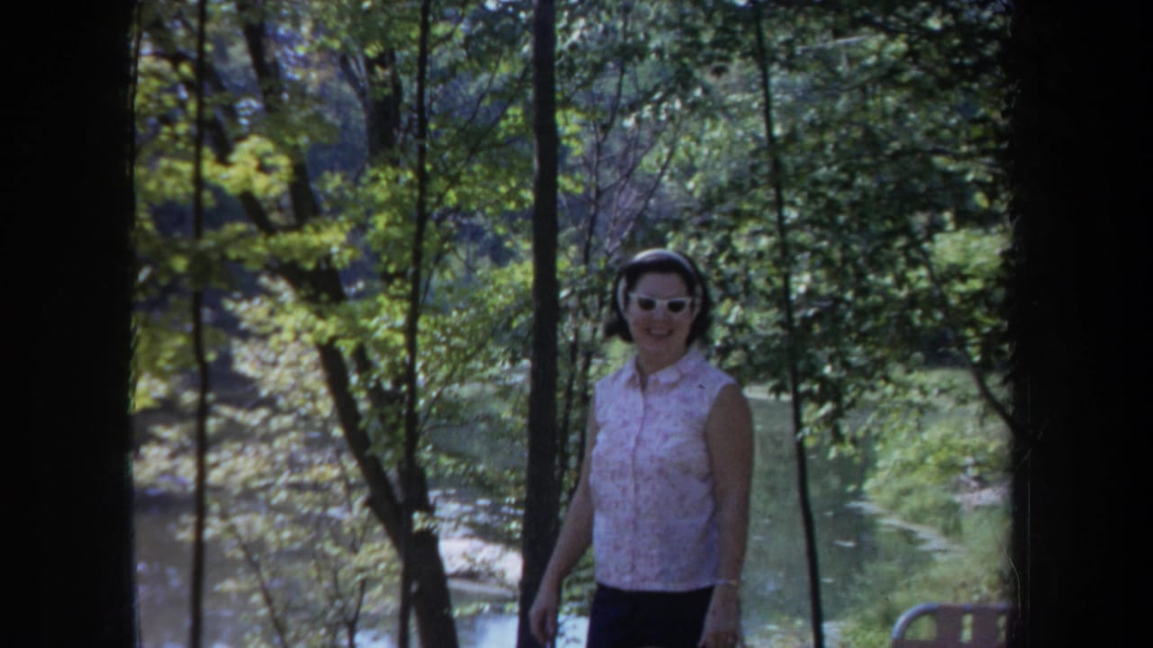 The woman smiles while walking through a wooded Michigan scene, with sunlit water or a pond visible behind her.