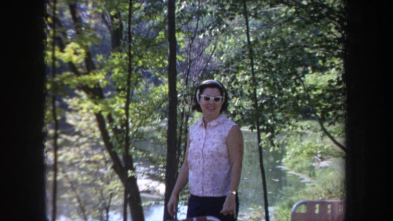 A 1968 Michigan home-movie frame shows the woman near trees and water, with outdoor seating partly visible at the edge.
