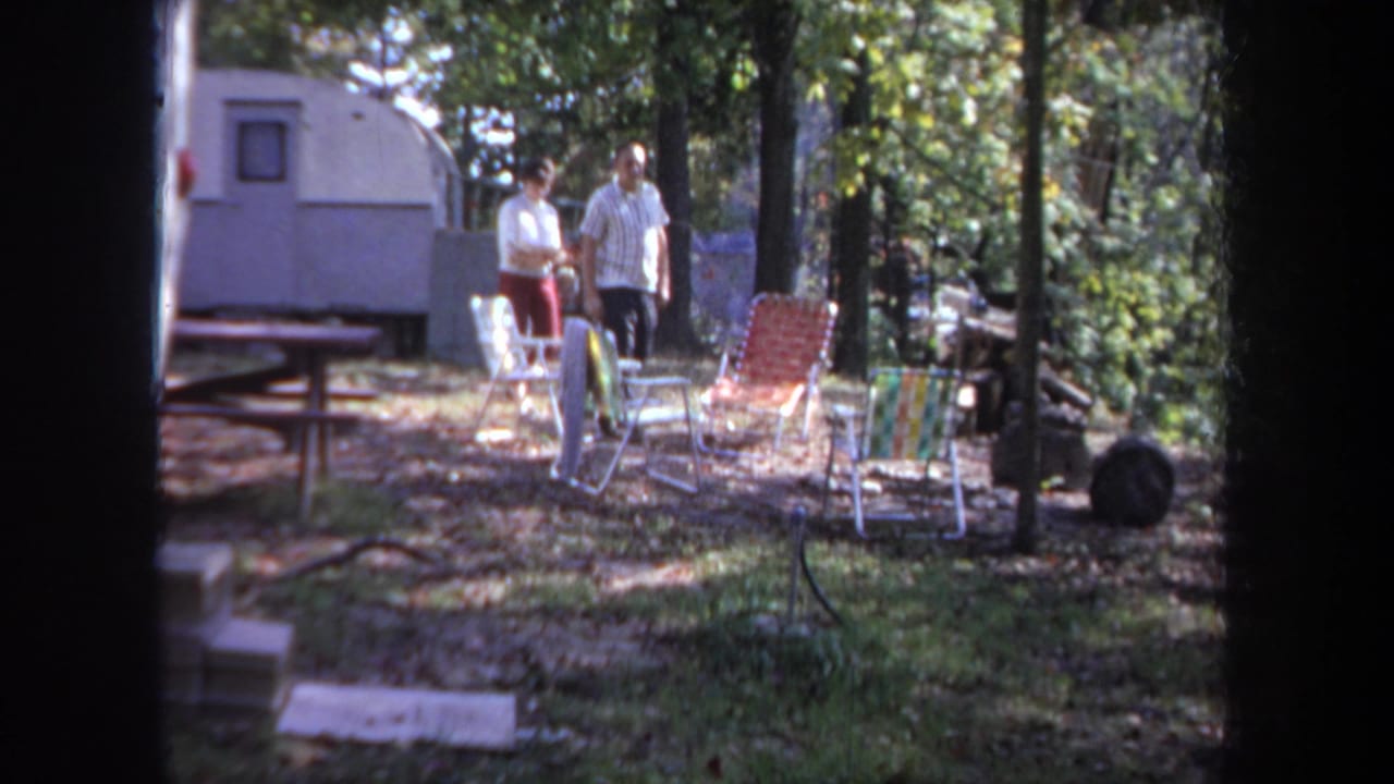 In 1968 Michigan home-movie footage, two adults walk past folding lawn chairs near a white camper trailer in a wooded campsite.