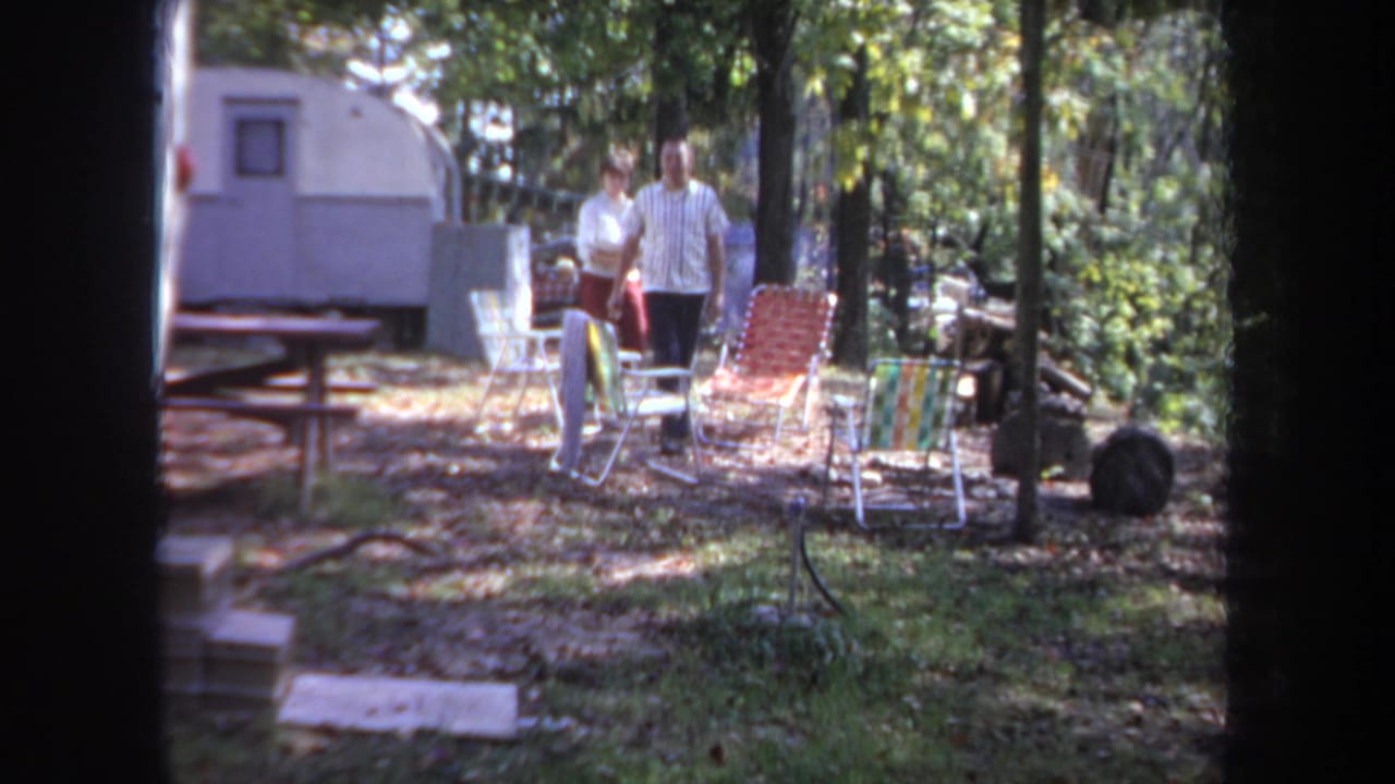 A man and woman walk toward the camera through a Michigan campsite, with folding chairs and a camper trailer visible behind them.