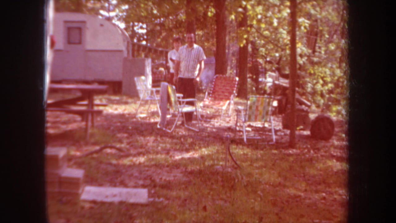 The adults appear among folding lawn chairs beside a white camper trailer, with warm color shift and dark 8mm frame edges.
