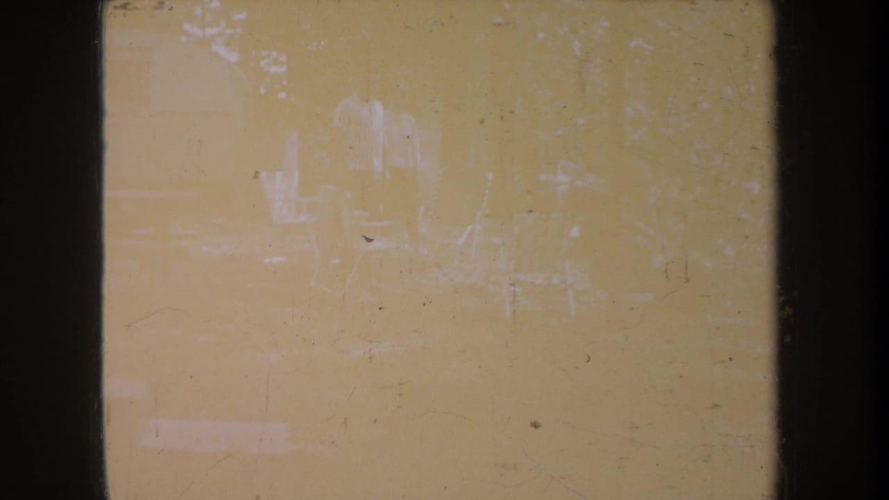 Faded 1968 Michigan 8mm frame with barely visible figures, chairs, trees, dust, scratches, and dark film borders.