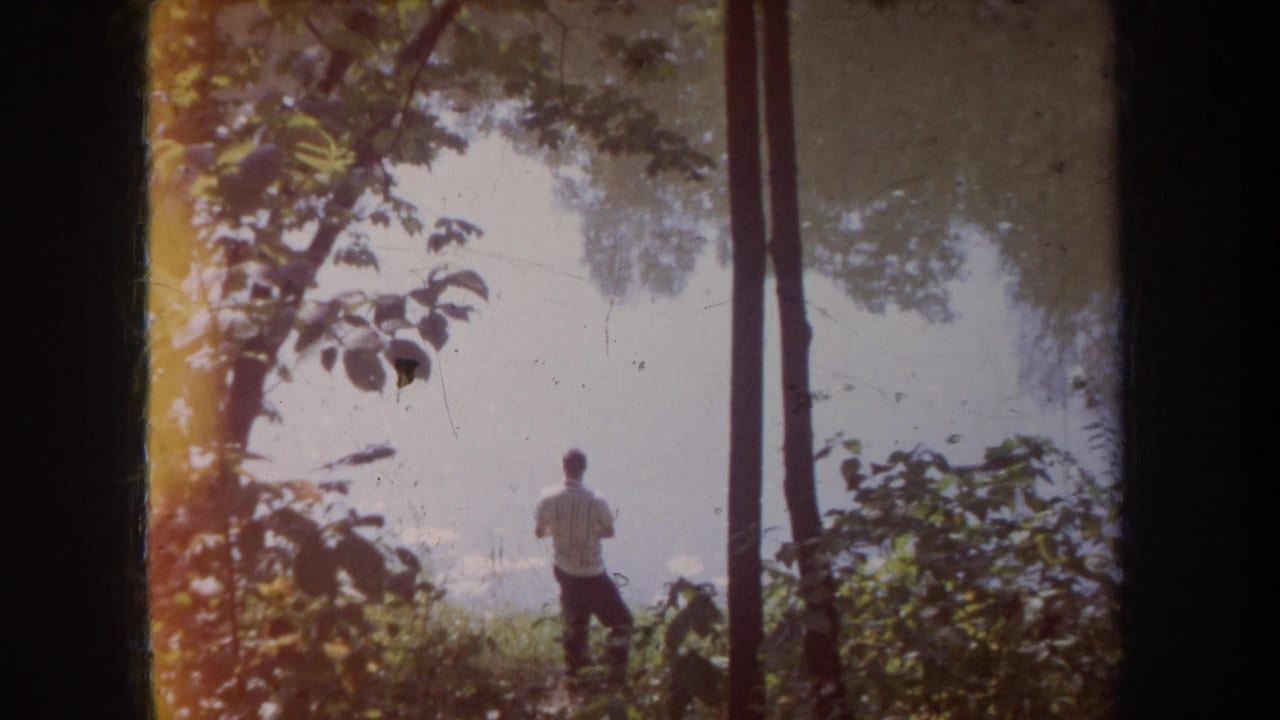 1968 Michigan 8mm frame showing a man in a striped white shirt near calm water, partly obscured by trees and leafy growth.
