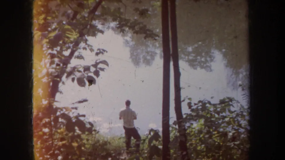 1968 Michigan 8mm frame showing a man in a striped white shirt near calm water, partly obscured by trees and leafy growth.