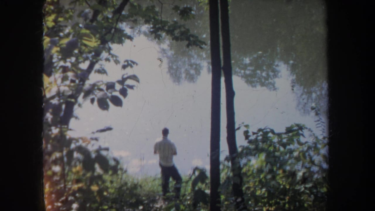 A man faces the water in a wooded Michigan setting, with soft 8mm grain and foliage framing the pond view.