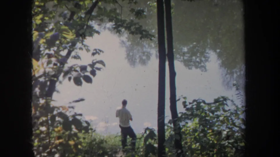 Michigan 8mm footage frame of a man at a quiet pond, seen through dark trunks and leafy branches with faded color and soft focus.