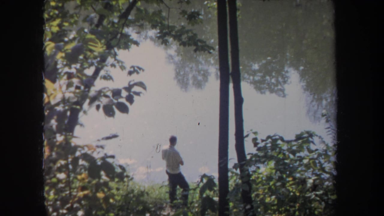 In 1968 Michigan home-movie footage, a man stands near a lake through a frame of leafy trees and trunks.