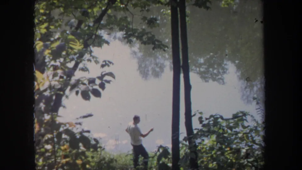 The lakeside view shows the man continuing a fishing motion among trees, brush, and bright water.