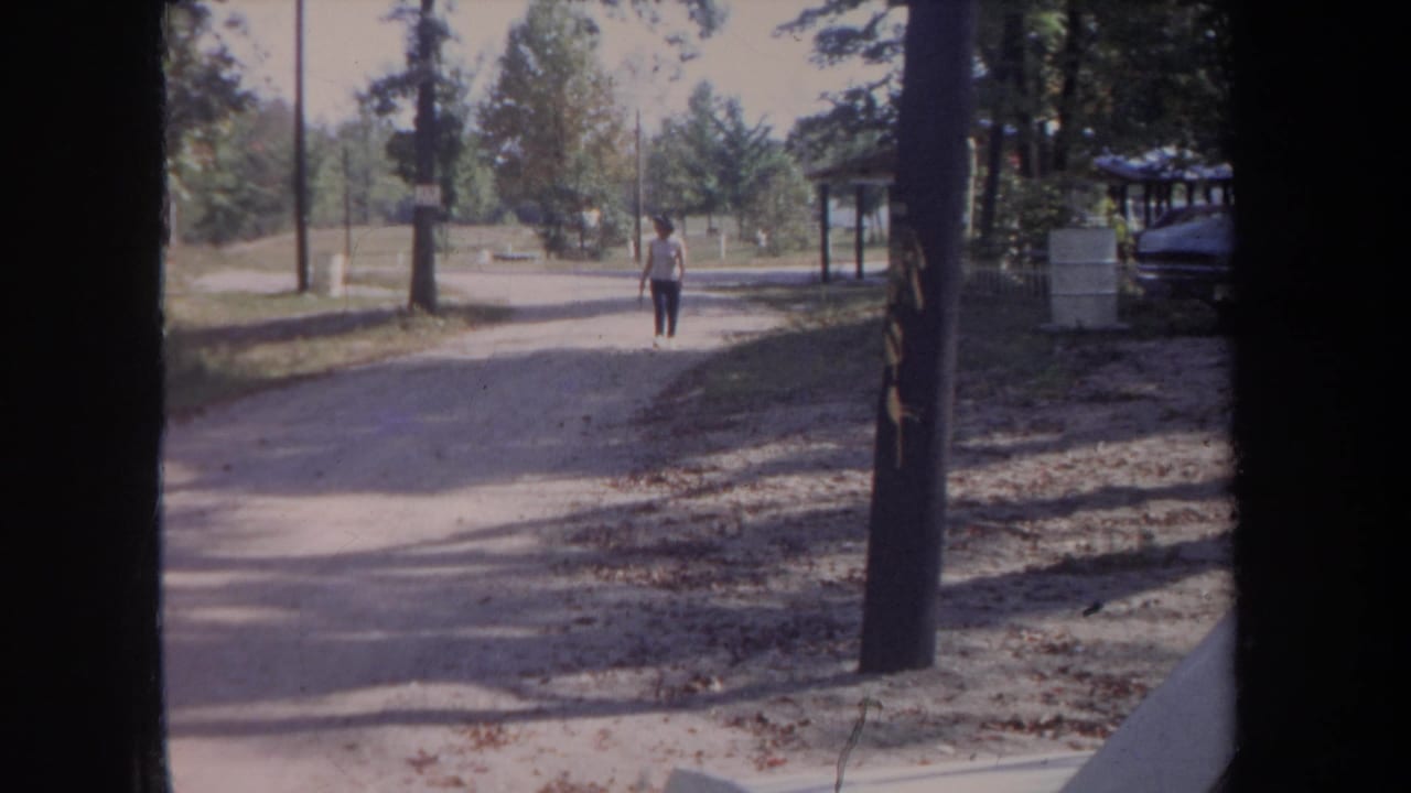 In 1968 Michigan home-movie footage, a woman in a white shirt walks toward the camera on a sunlit gravel path in a wooded park setting.