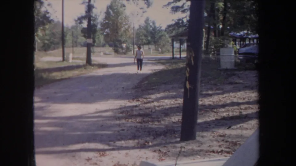 In 1968 Michigan home-movie footage, a woman in a white shirt walks toward the camera on a sunlit gravel path in a wooded park setting.