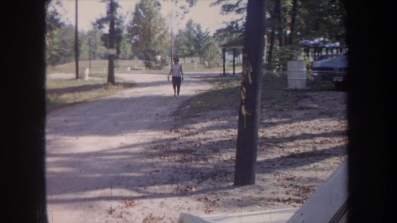 Woman continues down the gravel path with a parked car and shelter partly visible at right.