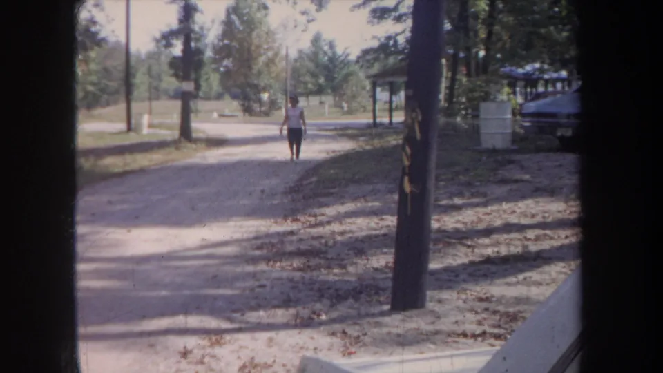 The woman walks closer along the wooded Michigan path, with a dark frame edge, parked car, and picnic shelter visible in the background.