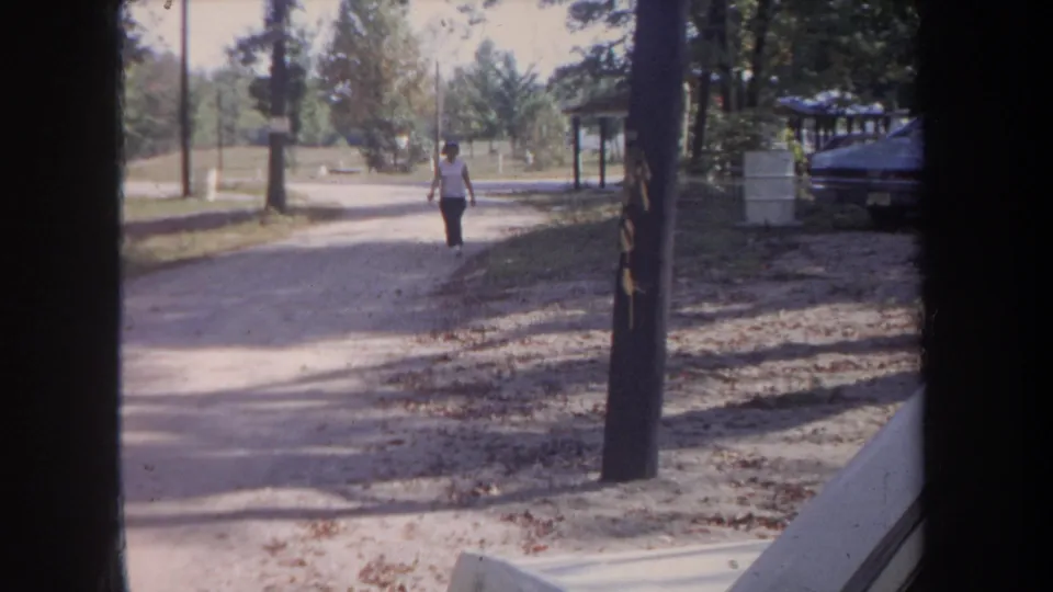 A woman in a white shirt and dark pants approaches on a curving gravel path, filmed through a dark foreground edge in 1968 Michigan.